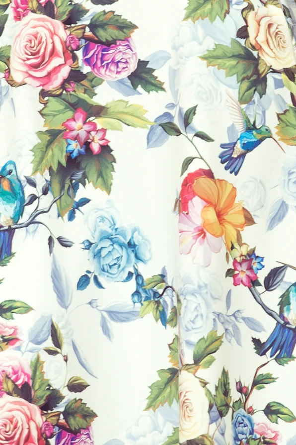 A vibrant maxi dress featuring a colorful floral pattern with roses, leaves, and hummingbirds on a light background. The design includes a flowing silhouette with a delicate ruffle detail, perfect for a summery, elegant look.