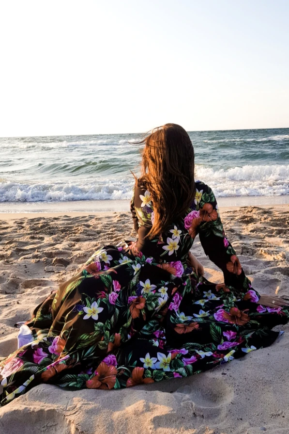 A woman sits on the beach wearing a flowing maxi dress adorned with vibrant floral patterns. The dress features a ruffled design, adding elegance to the relaxed seaside setting.
