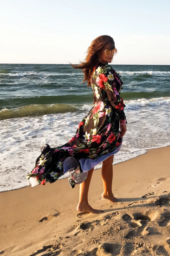 A woman is wearing a flowing maxi dress with a vibrant floral pattern, perfect for a beach setting. The dress features a delicate ruffle detail, adding elegance and movement as it catches the breeze.