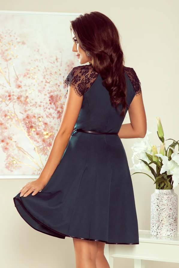 A woman is wearing an elegant dark teal dress with delicate lace cap sleeves and a flared skirt. The dress is complemented by a subtle belt at the waist, creating a sophisticated and feminine silhouette.