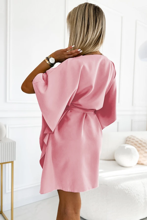 A woman is wearing a soft pink dress with wide sleeves and a belt that cinches at the waist, creating an elegant silhouette. The dress is made from a smooth fabric, perfect for both casual and formal occasions.