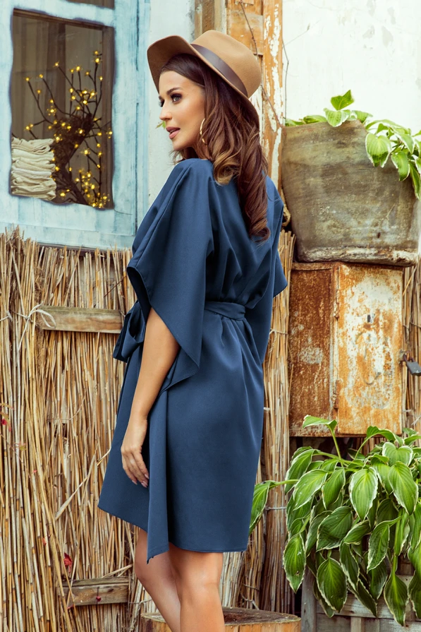 A woman is wearing a stylish, loose-fitting blue dress with wide sleeves and a belt that cinches at the waist. The dress is complemented by a brown hat, creating a chic and casual look.