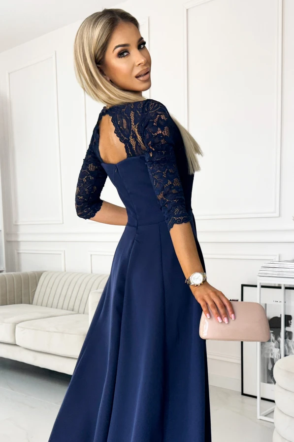 A woman is wearing an elegant navy blue dress with lace detailing on the sleeves and back. She holds a light pink clutch, standing in a sophisticated, modern interior.
