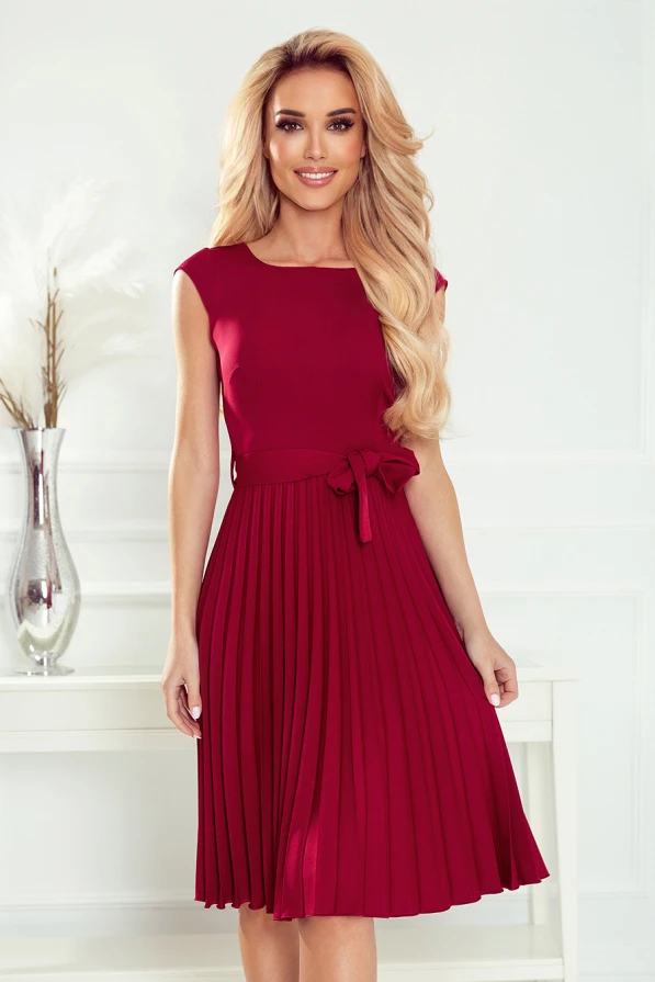 A woman is wearing a sleeveless, knee-length, pleated dress in a rich burgundy color, featuring a fitted bodice and a decorative bow at the waist. The elegant design is complemented by a simple, sophisticated backdrop with a vase of white feathers.