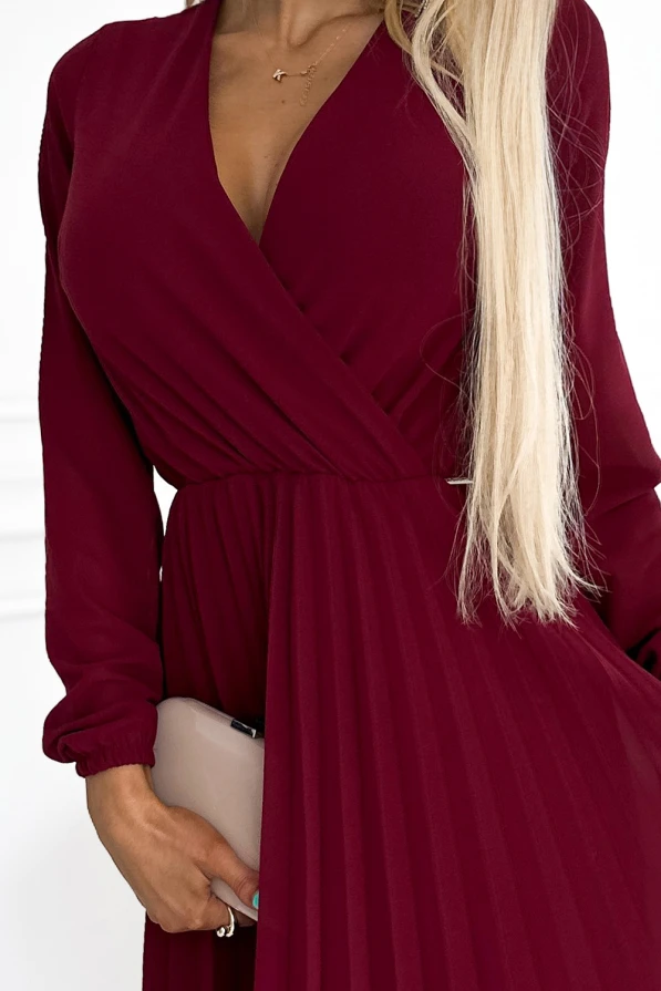 A woman is wearing a deep burgundy, pleated dress with long sleeves and a wrap-style V-neckline. She holds a beige clutch, complementing the elegant and sophisticated look of the outfit.
