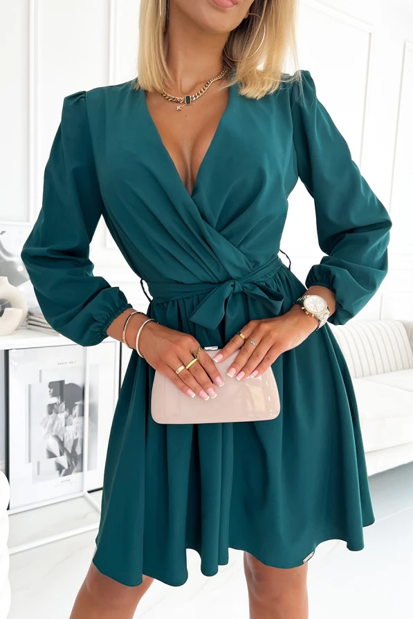 A woman is wearing an elegant teal dress with a deep V-neckline and long sleeves, featuring a tied waist that accentuates the silhouette. She accessorizes with a small, glossy clutch and layered jewelry, adding a touch of sophistication to the outfit.