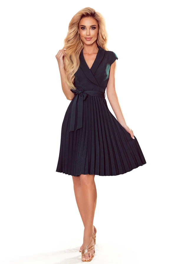 A woman is wearing an elegant, dark green pleated dress with a wrap-style top and a tied waist belt. The dress has cap sleeves and falls just above the knees, paired with gold sandals.