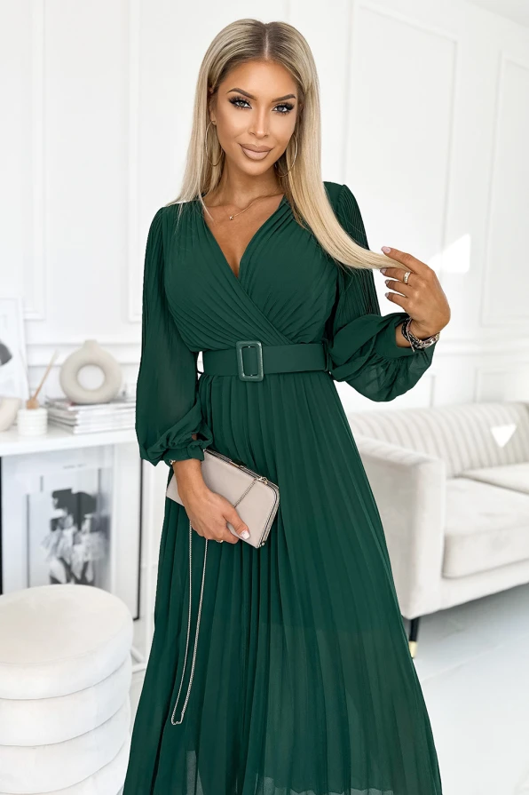 A woman is wearing an elegant, pleated green dress with long sleeves and a matching belt. She holds a beige clutch, standing in a stylish, modern interior.