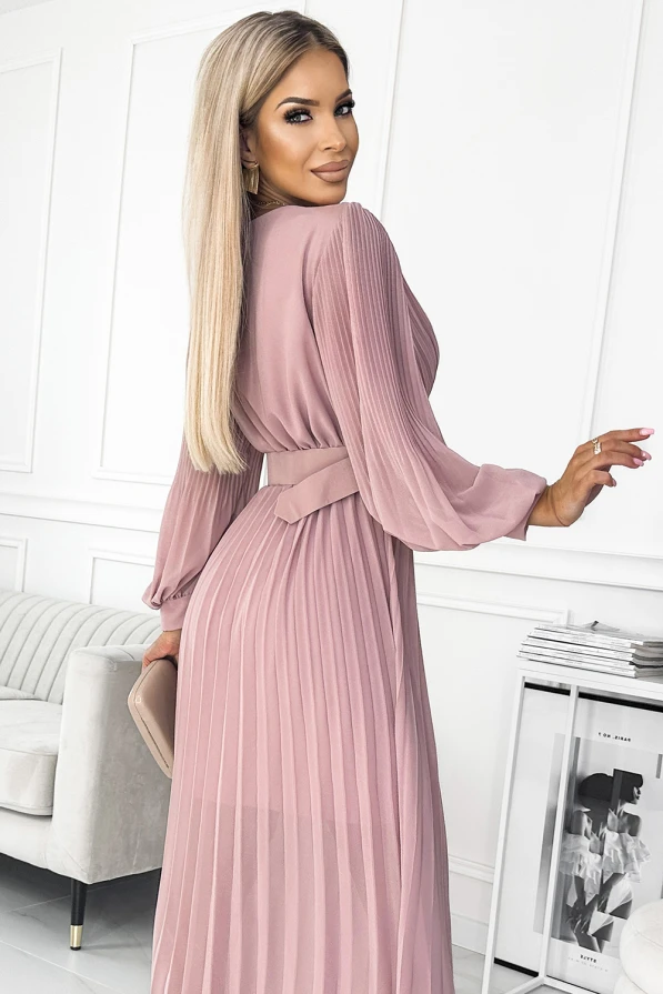 A pleated dress in a soft, dusty pink hue features long sleeves and a matching belt that accentuates the waist. The elegant design is complemented by a flowing skirt, creating a sophisticated and graceful silhouette.