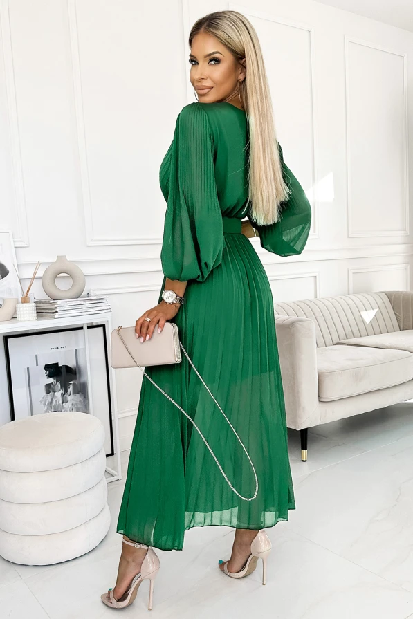 A woman is wearing a flowing, pleated green dress with long sleeves, perfect for elegant occasions. She accessorizes with a beige clutch and nude high heels, complementing the dress's sophisticated style.