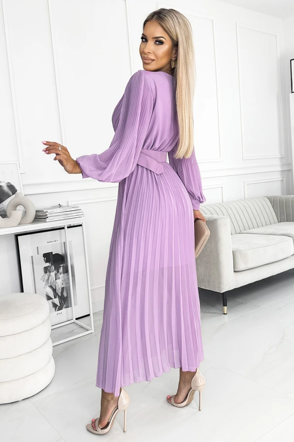 A woman is wearing a long, pleated lavender dress with a matching belt that accentuates the waist. The dress features long sleeves and a flowing, elegant silhouette, perfect for a sophisticated look.