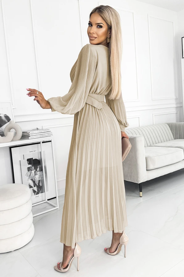 A woman is wearing a beige, pleated midi dress with long sleeves and a matching belt, standing in a modern, elegantly decorated room. The dress has a flowing, sophisticated design, perfect for both formal and casual occasions.