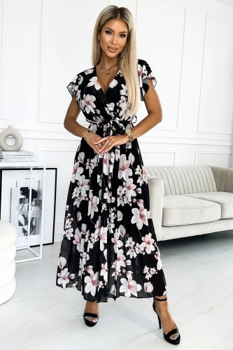 A woman is wearing a black pleated dress adorned with a floral pattern in shades of pink and white. The dress features short sleeves and a tied waist, creating an elegant and feminine silhouette.}