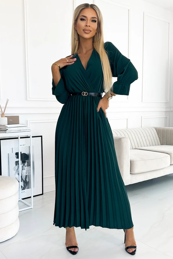 A woman is wearing an elegant, dark green pleated dress with a V-neckline and long sleeves, complemented by a stylish black belt. The dress flows gracefully to the floor, creating a sophisticated and timeless look.
