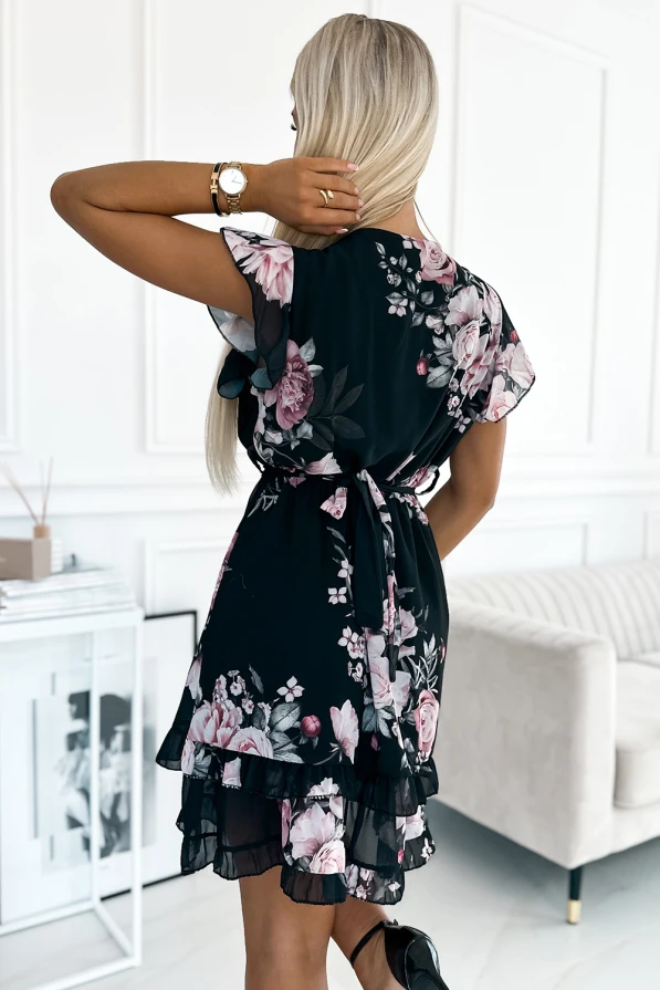 A woman is wearing a floral dress with a black base, featuring pink and white flowers and ruffled layers. The dress has short sleeves and a tie at the waist, creating a feminine and elegant silhouette.