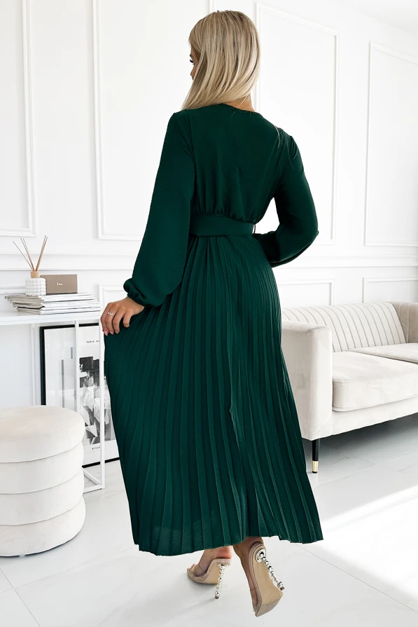 A woman is wearing a long, pleated dark green dress with long sleeves, creating an elegant and flowing silhouette. The dress is paired with stylish high heels, adding a touch of sophistication to the overall look.