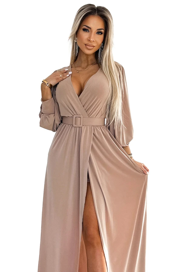 A long, elegant dress in a soft beige color features a deep V-neckline and a stylish belt at the waist. The flowing fabric and slit add a touch of sophistication and allure.